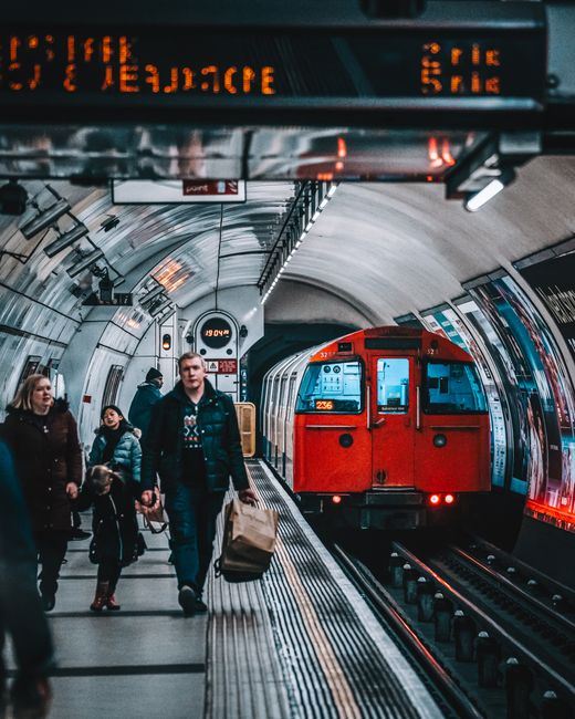 An underground London tube station platform with an orange-red train arriving or departing, showing the train's front window and lights. Several passengers, including a man holding a paper shopping bag and a woman with a child, stand on the platform near the train doors. The platform features a curved ceiling with metallic panels, digital signage displaying station information, and safety markings along the edge. Overhead lighting illuminates the scene, creating a well-lit environment suitable for home relocation or furniture transport logistics. This setting is part of the London Transport network, reflecting the typical environment encountered during city-based moving services. Man With a Van Barnet may assist with local moves requiring transit through such stations.