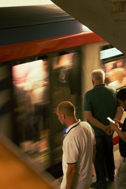 Two men stand on the platform of a home relocation or moving service, waiting to board a train at High Barnet tube station as part of a relocation process. One man, dressed in a white polo shirt with dark trim, looks down at his mobile device while facing the train, which is captured in motion blur, indicating the train’s departure or arrival. The second man, wearing a dark green shirt, stands with his back to the camera, observing the scene. The station's lighting is dim, with the train’s corridor illuminated and reflections visible on the train's windows, showing blurred figures inside. The platform surface is visible beneath their feet, and the scene reflects a common aspect of local moves involving transport logistics, highlighting the importance of timing and coordination in house removals serviced by companies like Man With a Van Barnet.