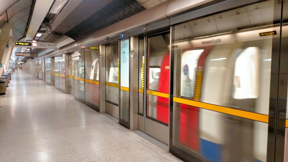 An underground London tube station platform with an orange-red train arriving or departing, showing the train's front window and lights. Several passengers, including a man holding a paper shopping bag and a woman with a child, stand on the platform near the train doors. The platform features a curved ceiling with metallic panels, digital signage displaying station information, and safety markings along the edge. Overhead lighting illuminates the scene, creating a well-lit environment suitable for home relocation or furniture transport logistics. This setting is part of the London Transport network, reflecting the typical environment encountered during city-based moving services. Man With a Van Barnet may assist with local moves requiring transit through such stations.