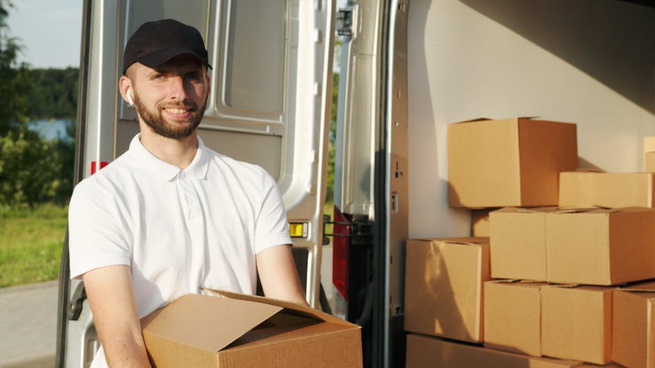 A man with a beard and wearing a black cap and white polo shirt is standing outside a white moving van, holding a cardboard box. The van's back doors are open, revealing a loading area with several st